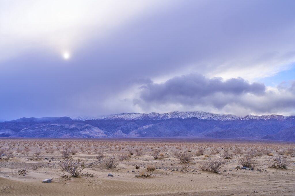 sandige Strauchlandschaft und lila blaue Berge im Hintergrund; die Sonne bemüht sich durch dichte Wolken