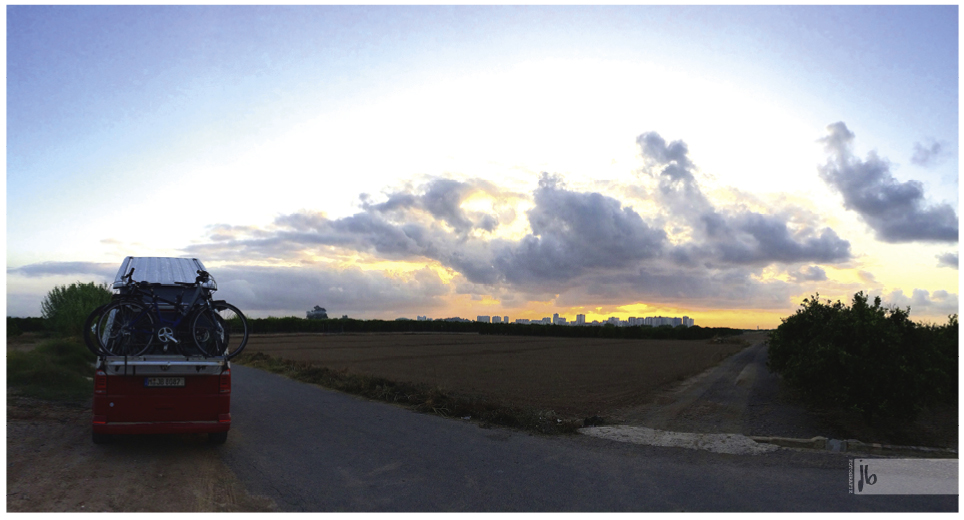 der rot weiße VW Bus bei Sonnenuntergang an einem Wegrand, die Stadt im Hintergrund, Wolken darüber