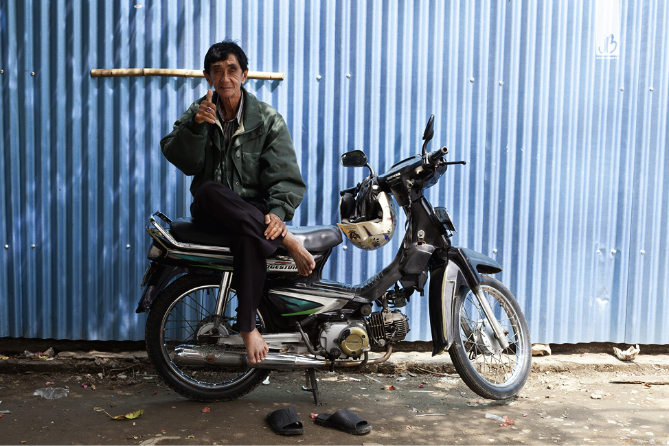 Indonesier sitzt auf seinem Moped und streckt den Daumen hoch mit Blick in die Kamera, hinter ihm eine blaue Wellblechwand