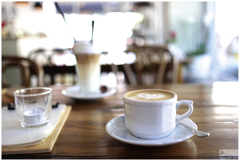 ein Cappuccino mit einem Milchherz und im Hintergrund verschwommen ein Glas Latte Macchiato sowie schemenhaft das Cafe selbst.