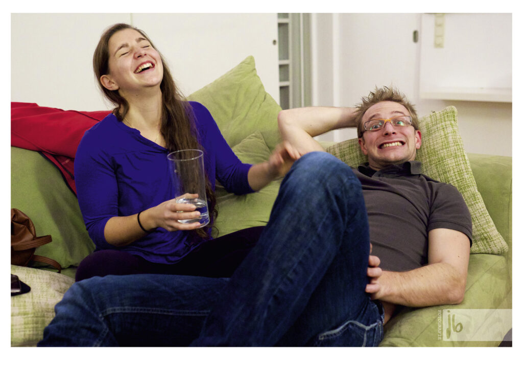 Ein Pärchen sitzt auf einer grünen Couch und Mona lacht herzlich mit einem Glas Wasser in der Hand während Beri in die Kamera grinst.