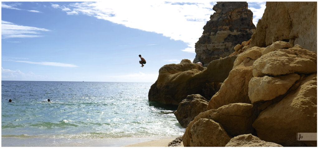 ein Junge springt mit angezogenen Beinen von einem Felsen ins Wasser der portugiesischen Algarve