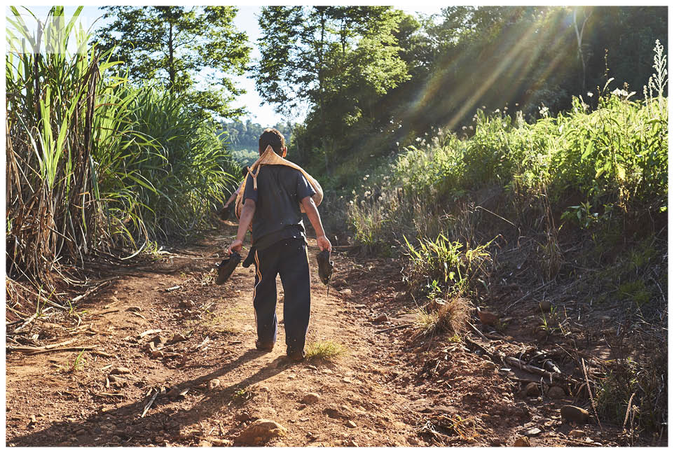 ein junge aus dem Guarani Dorf geht barfuß über einen Feldweg, seine Schuhe in der Hand und einen Sack mit Melonen um den Hals