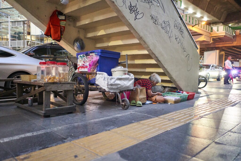 eine alte Dame unter einer Treppe in Bangkok geht einer Handarbeit nach, die sie verkauft