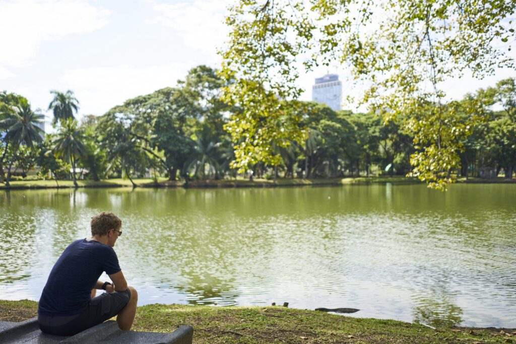 Ein Mann (Tom) sitzt auf einer Bank im Lumpini Park und beobachtet einen Leguan wie er aus dem Wasser kommt.