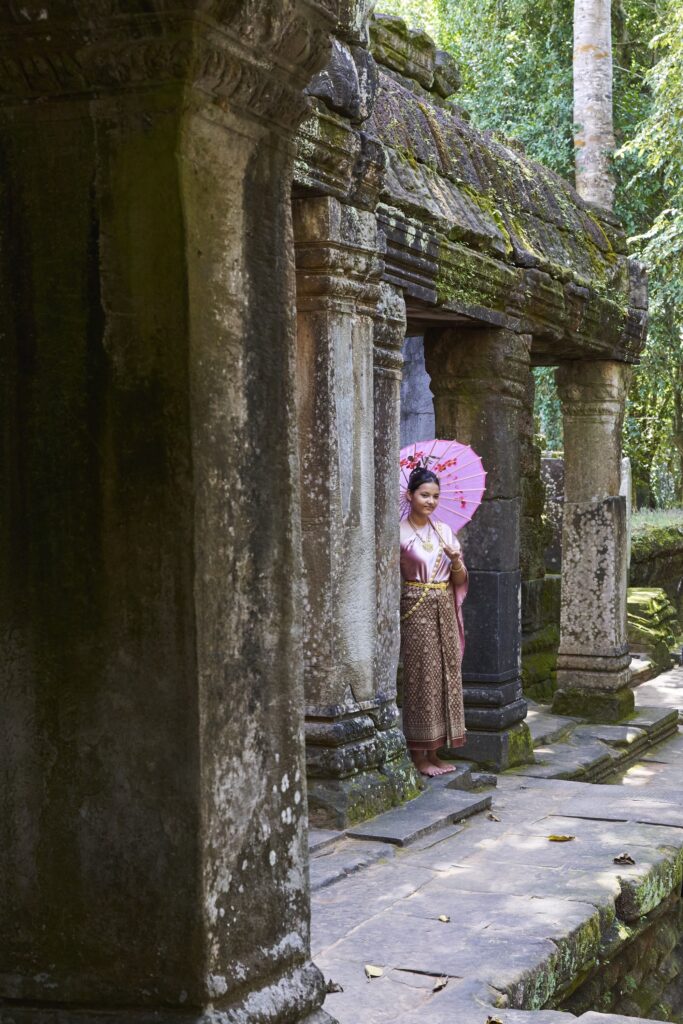 eine Frau in traditioneller Kleidung und einem Sonnenschirm, typisch für Asiatinnen steht in den Ruinen des Ta Prohm Tempels