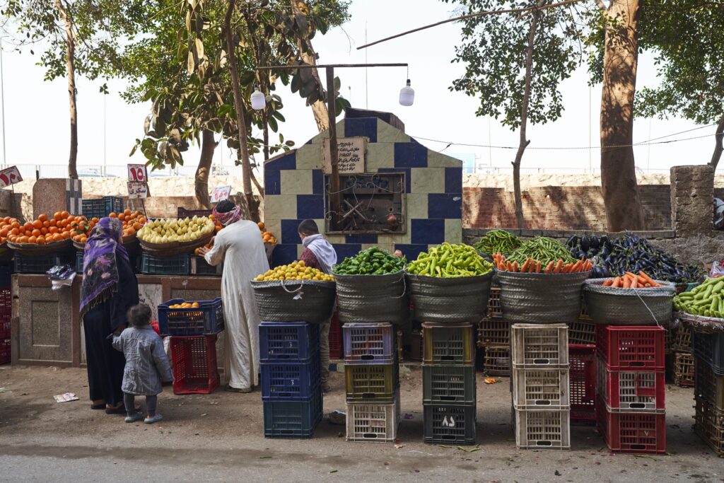 eine Frau mit Kopftuch hält ihr Kind an der Hand und begutachtet das Gemüse eines Standes in Kairo, der Mann bietet die Ware an.