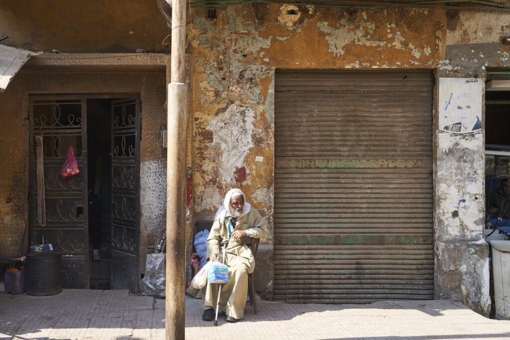 ein Mann sitzt mit Kopftuch und Gehstock auf einem Stuhl vor einem Garagentor in Kairo 