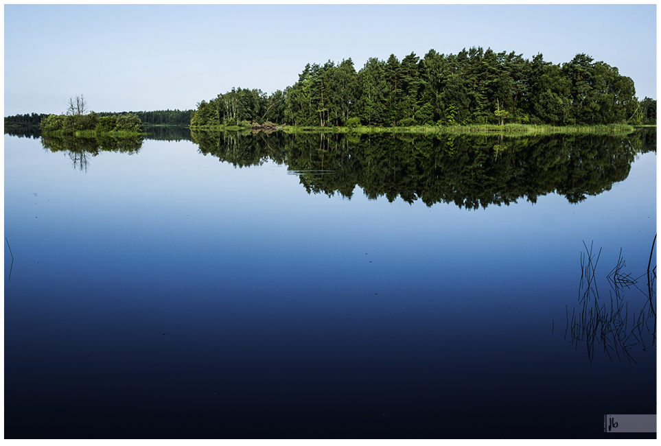 ein See in Schweden mit spiegelglatter Oberfläche und einer bewachsenen Insel von Bäumen in der Mitte, die sich fast ungestört spiegeln.