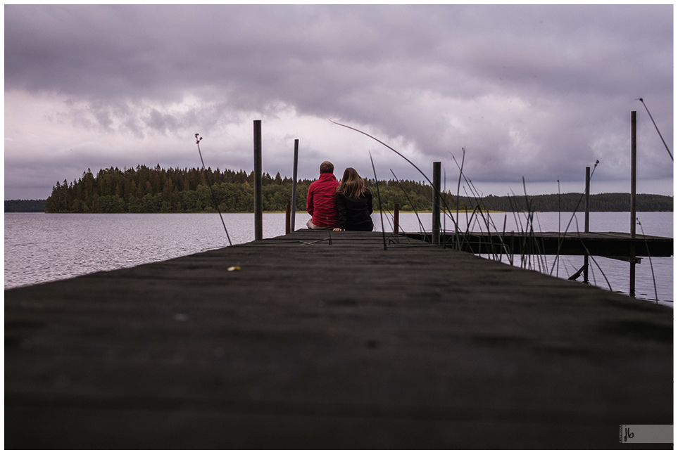 ein Pärchen von hinten, das auf einem Steg sitzt und auf einen See in Schweden blickt, der Himmel ist bewölkt, sie trägt ihre Haare offen und eine schwarze Jacke während er eine rote Jacke trägt