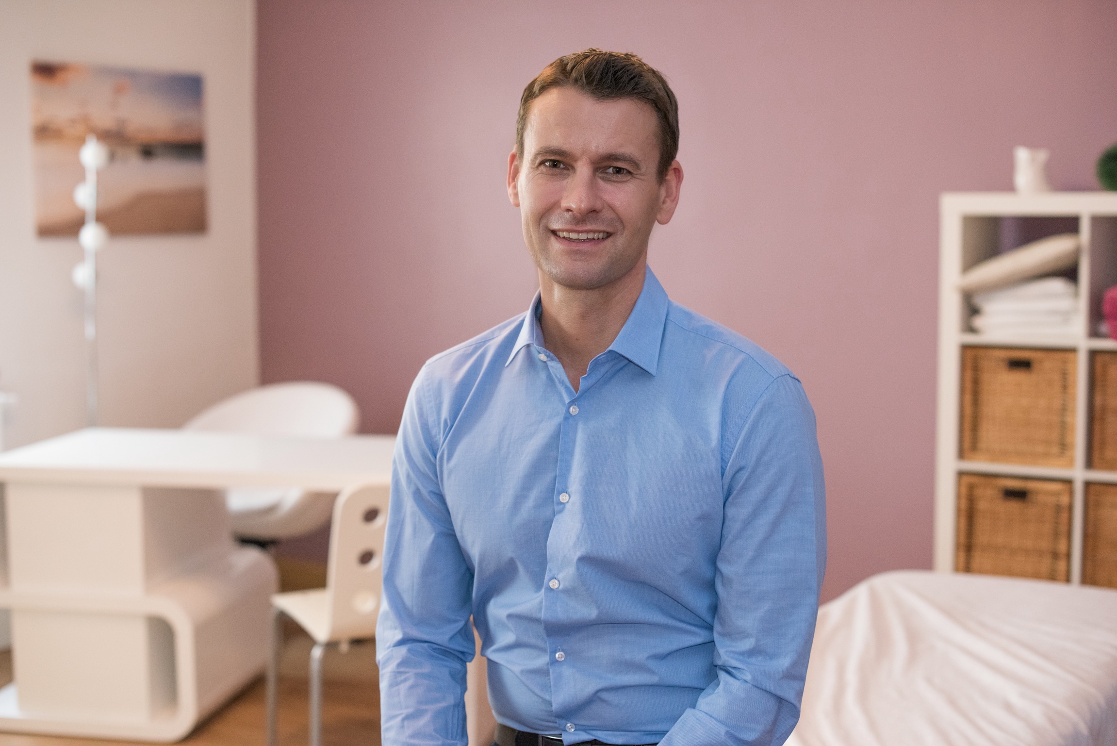 Stefan Rieth, Osteopath sitzt in einem blauen Hemd in seiner Praxis und lächelt in die Kamera