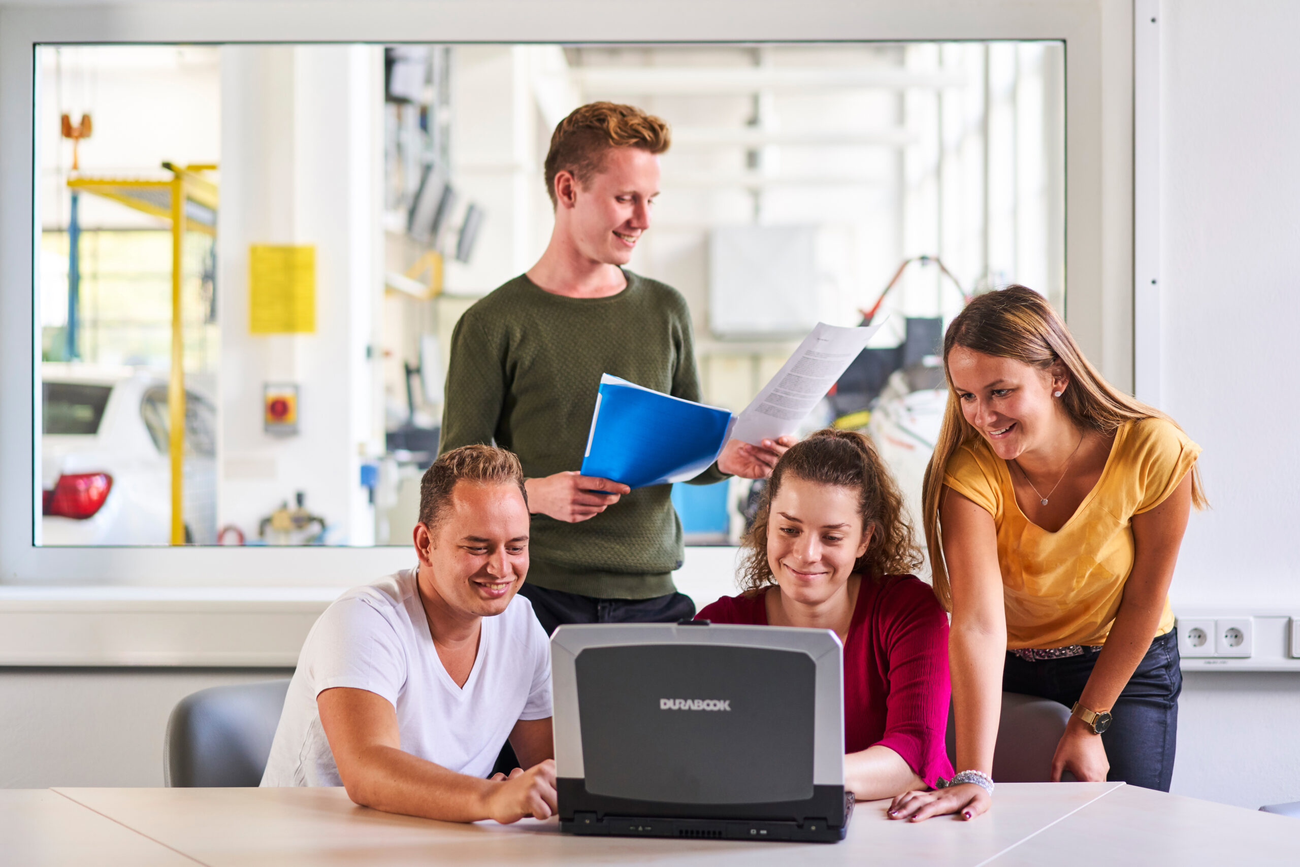 dual Studierende beugen sich über einen Laptop, einer hält im Hintergrund Zettel in der Hand, dahinter das Glasfenster zum Labor